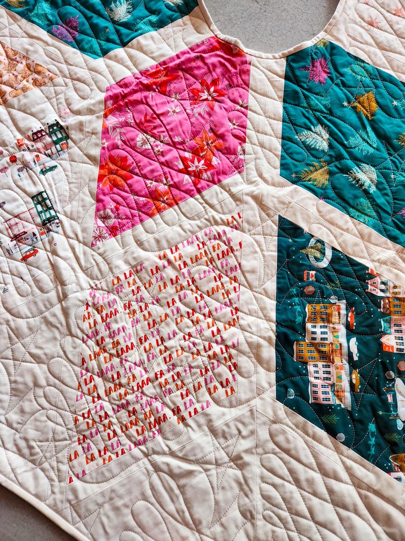 May include: A quilted textile featuring a patchwork design with various geometric shapes. The quilt showcases vibrant colors, including pink floral patterns, teal, and a cream-colored background. The text "LA LA LA" is visible on one of the patches.