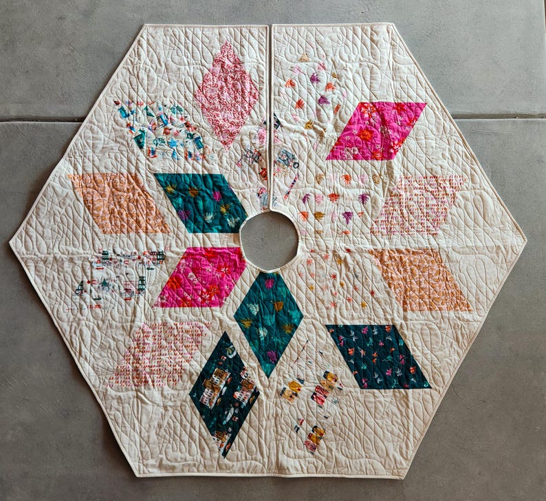 May include: A quilted, cream-colored tree skirt with a hexagonal shape. It features diamond-shaped patches in pink, teal, and peach, each with floral patterns. The skirt has a central hole and is stitched with a quilted design.