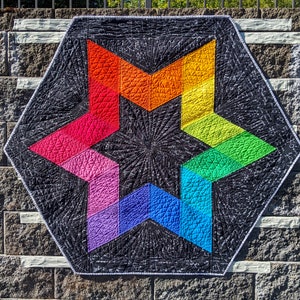 May include: A handmade, hexagonal quilt featuring a vibrant rainbow star design on a black background. The star is composed of segments in red, orange, yellow, green, blue, and purple. The quilt has intricate stitching and a white border.