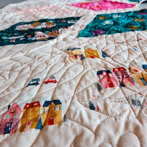 May include: A quilted blanket with a cream-colored background and colorful patches. The patches feature illustrations of houses, cars, and trees. The quilt has a wavy, stitched pattern throughout.