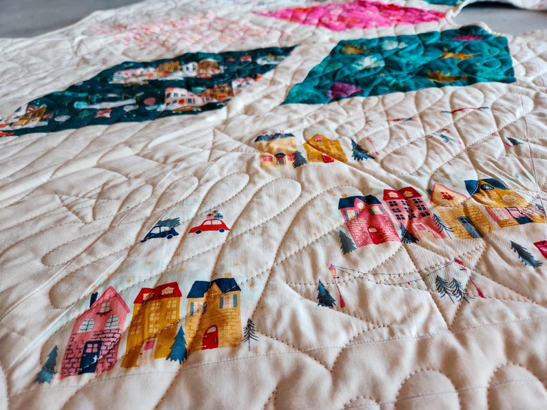 May include: A quilted blanket with a cream-colored background and colorful patches. The patches feature illustrations of houses, cars, and trees. The quilt has a wavy, stitched pattern throughout.