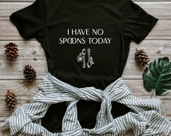 May include: Black t-shirt with the text "I HAVE NO SPOONS TODAY" in white. The shirt features a graphic of two spoons and a flower. A striped shirt is tied around the waist. The shirt is on a wooden surface with pinecones and a leaf.