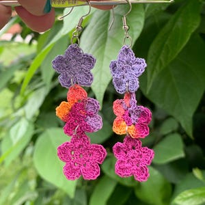 Puede incluir: Un par de pendientes de flores de ganchillo hechos a mano. Cada pendiente presenta tres flores en tonos morados, naranjas y rosas. Los pendientes están suspendidos de ganchos plateados. El fondo es un exuberante follaje verde.