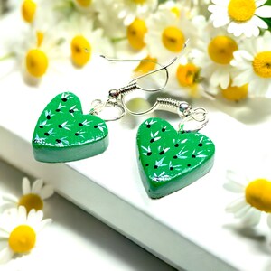 Könnte beinhalten: Ein Paar grüne herzförmige Ohrringe mit einem weißen Blumenmuster. Die Ohrringe sind aus Holz und haben silberne Haken.