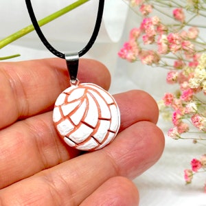 May include: A white and peach-colored concha pendant necklace with a black cord. The pendant is a detailed replica of a traditional Mexican sweet bread.