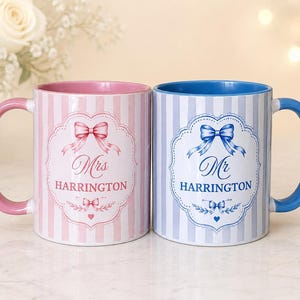 May include: Two ceramic mugs featuring vertical stripes and a central decorative frame. The pink mug displays the text Mrs HARRINGTON with a bow graphic, while the blue mug displays the text Mr HARRINGTON with a matching bow graphic.