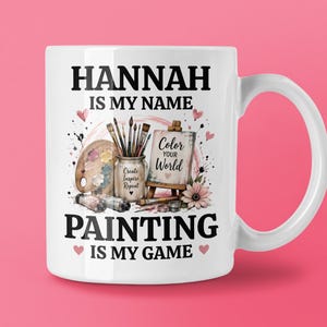 May include: A white ceramic mug featuring the black text HANNAH IS MY NAME PAINTING IS MY GAME. The graphic includes a paint palette, brushes in a jar labelled Create Inspire Repeat, and a small easel with the text Color Your World.
