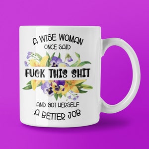 May include: White ceramic mug featuring the text "A WISE WOMAN ONCE SAID FUCK THIS SHIT AND GOT HERSELF A BETTER JOB" in black, with a floral arrangement of yellow lilies and purple pansies. The mug has a curved handle.
