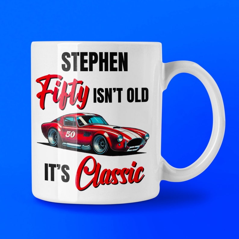 以下が含まれることがあります： 白いセラミックマグカップ。赤いスポーツカーのグラフィックと「STEPHEN Fifty ISN'T OLD IT'S Classic」の文字。車は赤と白のストライプで、ナンバー50。背景は青色。