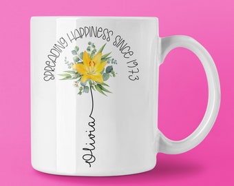 Puede incluir: Taza de cerámica blanca con un diseño floral y el texto "Spreading Happiness Since 1973" en negro. Una flor amarilla con hojas verdes y el nombre "Olivia" en escritura cursiva aparecen en la taza.