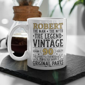 May include: White ceramic mug with the text "ROBERT THE MAN THE MYTH THE LEGEND VINTAGE 90" in gold and black. The mug has a curved handle and is on a black surface.
