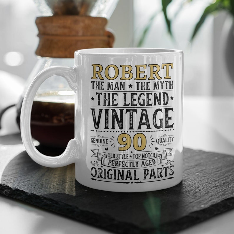 May include: White ceramic mug with the text "ROBERT THE MAN THE MYTH THE LEGEND VINTAGE 90" in gold and black. The mug has a curved handle and is on a black surface.