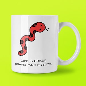 May include: White ceramic mug with a red cartoon snake and black spots. The mug has the text "Life is great, snakes make it better" in black. The mug has a white handle and is set against a bright green background.
