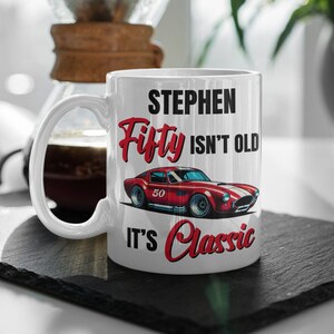 以下が含まれることがあります： 「STEPHEN Fifty ISN'T OLD IT'S Classic」の文字が黒と赤でプリントされた白いセラミックマグ。50の数字が入った赤いスポーツカーのグラフィックも特徴です。マグは黒い表面に置かれています。