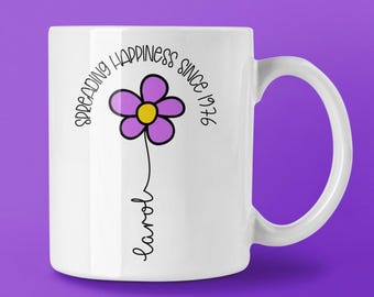 Puede incluir: Taza de cerámica blanca con un diseño de flor morada y el texto "SPREADING HAPPINESS SINCE 1976". El nombre "Carol" está escrito debajo de la flor en cursiva. La taza tiene un asa curva y está sobre un fondo morado.