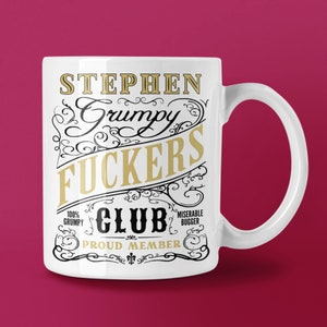 May include: White ceramic mug with the words "STEPHEN Grumpy FUCKERS CLUB" in gold and black lettering. The mug also includes the phrases "100% Grumpy", "Miserable Bugger", and "PROUD MEMBER". The mug has a curved handle and is set against a maroon background.