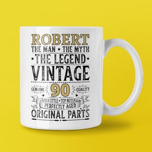 May include: White ceramic mug with the text "ROBERT THE MAN THE MYTH THE LEGEND VINTAGE 90 QUALITY" in gold and black lettering. The mug has a handle and is set against a yellow background.
