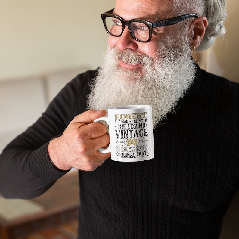 May include: White ceramic mug with the text "Robert The Man The Myth The Legend Vintage 90 Genuine Original Parts". The mug is held by a person wearing black glasses and a black sweater.