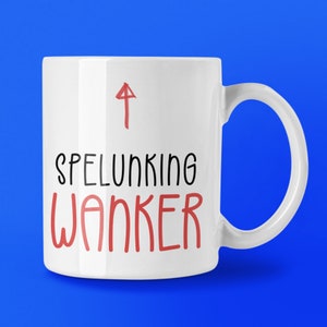 May include: White ceramic mug with the words "SPELUNKING WANKER" in black and red text, with a red arrow pointing upwards. The mug has a curved handle and is set against a blue background.