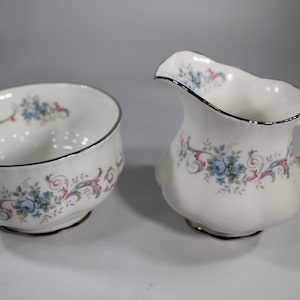 Könnte beinhalten: Ein weißes Porzellan-Zucker- und Milchkännchen-Set mit einem Blumenmuster in Blau-, Rosa- und Grautönen. Das Set hat einen silbernen Rand.