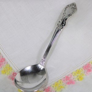 Puede incluir: Una cuchara de servir de plata con un mango adornado y un cuenco redondo. La cuchara está sobre una servilleta de lino blanca con un borde de ganchillo amarillo y rosa.