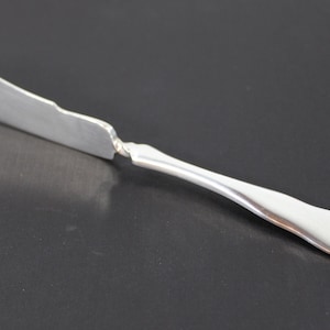 Puede incluir: Un cuchillo de mantequilla de plata con un mango decorativo. El mango tiene un borde festoneado y un diseño floral.
