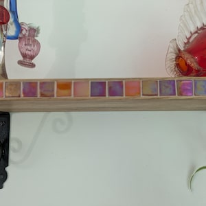 Op de afbeelding: Een houten wandplank met een voorrand versierd met een rij iriserende vierkante tegels in roze, oranje en paarse tinten. De plank wordt ondersteund door twee zwarte metalen beugels met kruldetails en bevat glazen ornamenten.