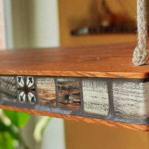 Op de afbeelding: Een houten plank met een moza&iuml;ekrand. De tegels zijn in tinten bruin, zwart en wit, met geometrische patronen. De plank is aan het plafond gehangen met een touw.