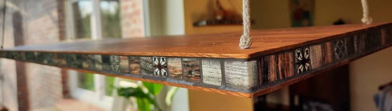 Op de afbeelding: Een houten plank met een moza&iuml;ekrand. De tegels zijn in tinten bruin, zwart en wit, met geometrische patronen. De plank is aan het plafond gehangen met een touw.