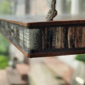 Op de afbeelding: Een houten plank met een decoratieve tegelrand. De tegels zijn bruin, wit en zwart met een geometrisch patroon. De plank hangt aan een touw.