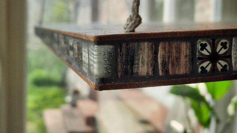 Op de afbeelding: Een houten plank met een decoratieve tegelrand. De tegels zijn bruin, wit en zwart met een geometrisch patroon. De plank hangt aan een touw.