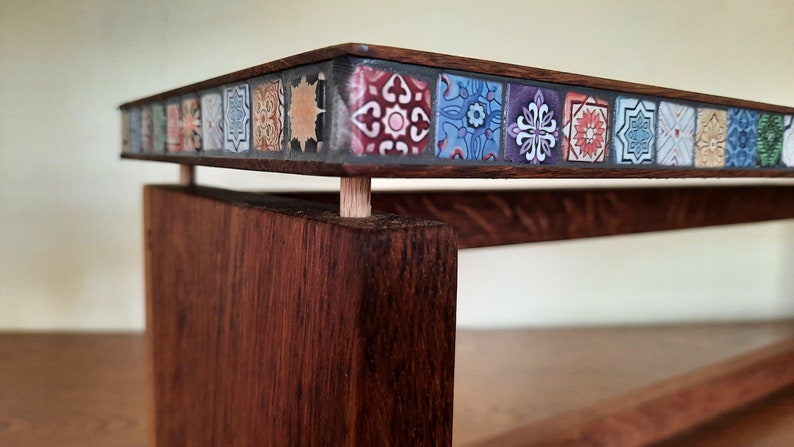 K&ouml;nnte beinhalten: Ein Holztisch mit einer Tischplatte aus bunten Mosaikfliesen. Der Tisch hat ein schlichtes Design mit einem einzigen Regal.