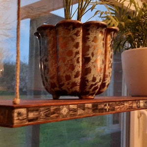 Op de afbeelding: Een houten hangende plank met een moza&iuml;ek tegelrand. Twee planten in potten staan op de plank, een in een witte pot en de andere in een gouden en witte pot.