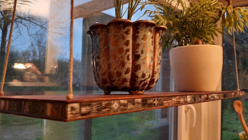 Op de afbeelding: Een houten hangende plank met een moza&iuml;ek tegelrand. Twee planten in potten staan op de plank, een in een witte pot en de andere in een gouden en witte pot.