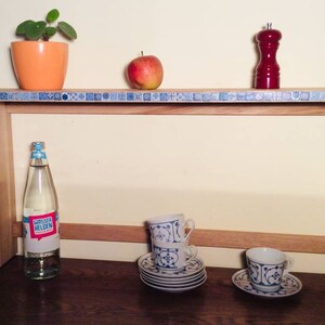 K&ouml;nnte beinhalten: Ein Holzregal mit einem blauen und wei&szlig;en Fliesenrand. Das Regal h&auml;lt eine Pflanze in einem orangenen Topf, einen roten Pfefferstreuer, einen Apfel, eine Wasserflasche und zwei Sets Teetassen und Untertassen.