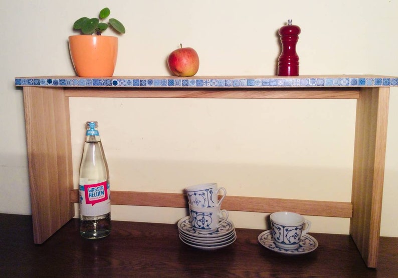 K&ouml;nnte beinhalten: Ein Holzregal mit einem blauen und wei&szlig;en Fliesenrand. Das Regal h&auml;lt eine Pflanze in einem orangenen Topf, einen roten Pfefferstreuer, einen Apfel, eine Wasserflasche und zwei Sets Teetassen und Untertassen.