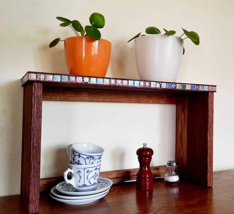 K&ouml;nnte beinhalten: Ein Holzregal mit einem dekorativen Fliesenrand, auf dem zwei Topfpflanzen stehen, eine mit einem orangen und eine mit einem wei&szlig;en Topf, sowie drei wei&szlig;e Teetassen mit blauen Blumendesigns. Das Regal steht auf einer Holzoberfl&auml;che.