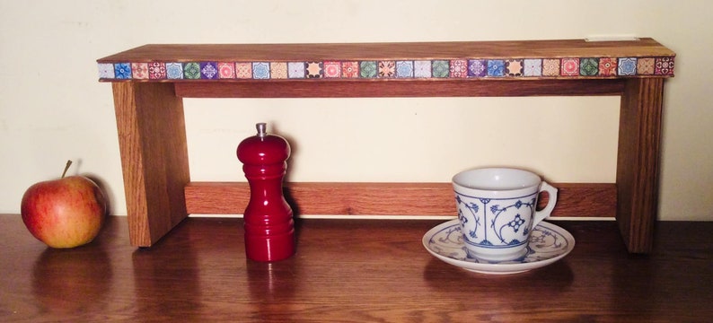 K&ouml;nnte beinhalten: Ein Holzregal mit einem dekorativen Fliesenrand, einem roten Pfefferm&uuml;hle, einer wei&szlig;en Teetasse mit blauem Blumendesign und einem roten Apfel auf einer Holzoberfl&auml;che.