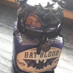 Può includere: Un piccolo barattolo di vetro con un liquido viola al suo interno. Il barattolo ha un coperchio nero con un design a forma di pipistrello e un ciondolo di metallo che dice "31" appeso ad esso. L'etichetta sul barattolo dice "Sangue di pipistrello" e "100% biologico".