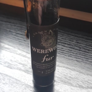 Può includere: Una piccola bottiglia di vetro con un'etichetta nera che recita "LONDON FINE WEREWOLF fur" e un sigillo di cera dorato sulla parte superiore. La bottiglia è appoggiata su una superficie di legno.