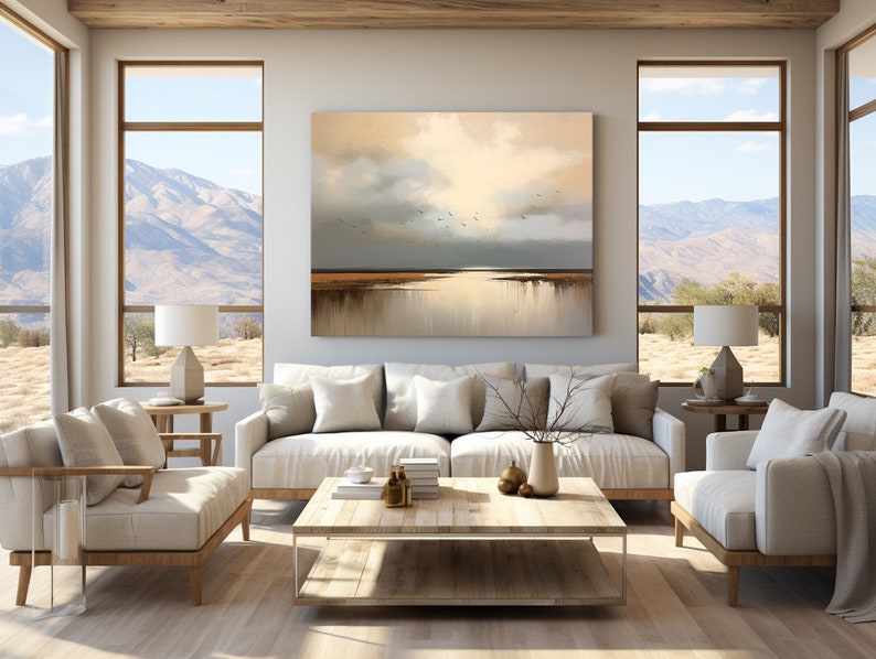 May include: A living room interior featuring a large abstract painting depicting a landscape with a cloudy sky and a body of water. The painting is hung on a wall with a white sofa, two armchairs, a coffee table, and two lamps. The room has large windows offering a view of mountains and a desert landscape.