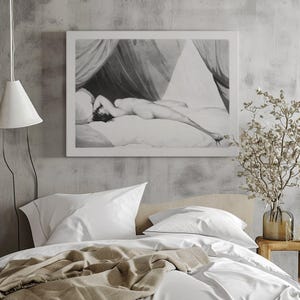 Peut inclure: Un cadre noir et blanc d'une figure allong&eacute;e sur un lit, mont&eacute; sur un mur gris. Le lit est fait de draps blancs et d'une couverture beige. Une lampe blanche est suspendue au plafond et un vase de fleurs s&eacute;ch&eacute;es est pos&eacute; sur une table en bois.