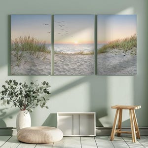 Könnte beinhalten: Ein dreiteiliger Leinwanddruck, der eine ruhige Strandszene zeigt. Das Kunstwerk zeigt einen Sandstrand, Meereswellen und einen Sonnenuntergangshimmel. Auch Seegras und Vögel sind zu sehen, was eine beruhigende Küstenästhetik schafft.