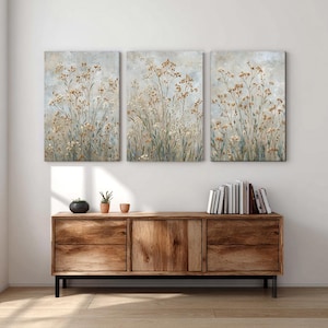 May include: A set of three canvas artworks depicting a field of tall, dried flowers in beige, brown, and green against a light blue-grey backdrop. The paintings are above a wooden cabinet with drawers and doors.