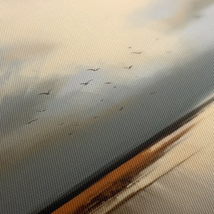 May include: A close-up of a canvas painting depicting a flock of birds flying in formation against a cloudy sky. The sky is a soft blue and white, with hints of orange and yellow. The birds are black silhouettes against the sky.