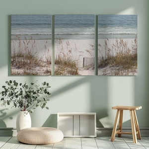 May include: A triptych canvas print depicting a serene beach scene. The artwork features a sandy beach with dune grass and a wooden fence, leading to the ocean with gentle waves. The color palette includes shades of blue, beige, and brown.