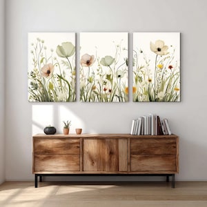 May include: A triptych of floral canvas prints showcasing watercolour-style flowers in pastel hues. The artwork depicts wildflowers and grasses on a cream backdrop. The prints are displayed above a wooden cabinet.