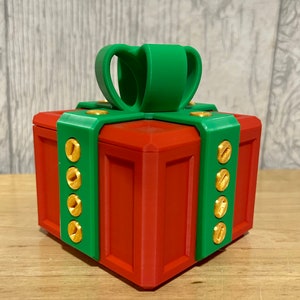 May include: A red and green gift box with a gold bow and gold accents. The box is made of plastic and has a hinged lid.