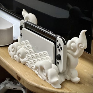 May include: White 3D printed stand shaped like clouds and dragons holding a Nintendo Switch console. The console is white with black buttons.