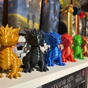 May include: A collection of seven brightly coloured dragon figurines, 3D-printed and arranged on a shelf. The dragons are in gold, black, blue, red, green, bronze, and orange. Books are visible in the background.